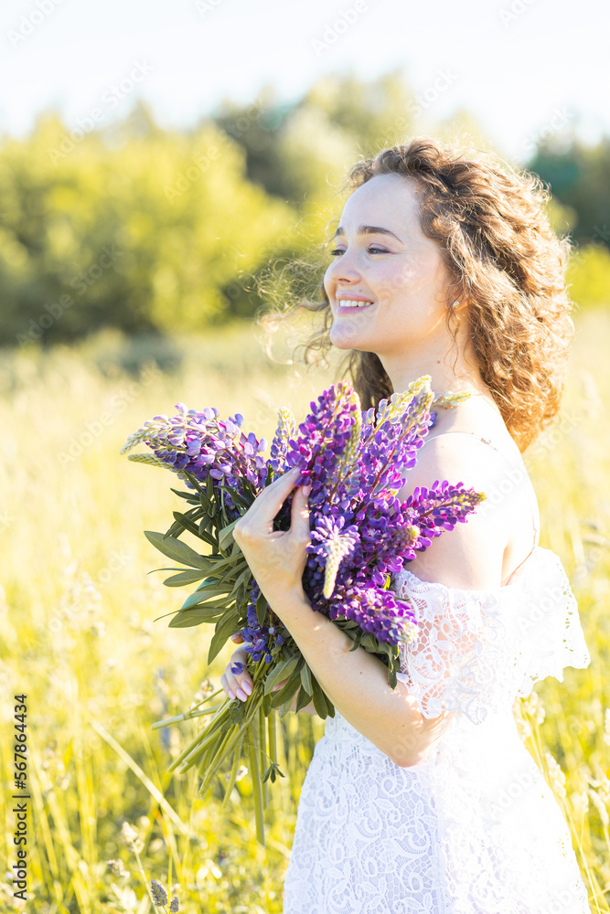 Fototapeta premium Beautiful young girl in a white dress, straw hat, with picnic and bouquet of purple wild flowers on a meadow. Summertime, golden hour, sunset gathering plants for handmade organic cosmetic production