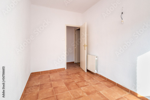 An empty spacious room with old square worn caramel colored floor tiles and a radiator on the wall  with an open door  white faded walls and ceiling. The wires for the lamp stick out in the wall.
