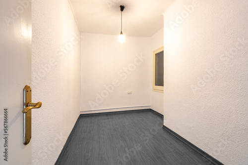 Entrance to an empty spacious unrenovated bedroom with cream walls and old black hardwood flooring. A lamp without a shade hangs from the ceiling, dimly illuminating the room. photo