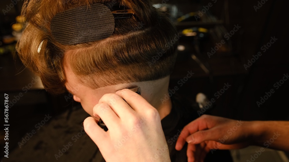 Fototapeta premium men's hairstyling and haircutting with barber shaving machine, hair cut for male. Hairdresser service with shaver in a modern barbershop in a dark key lightning with warm light side view