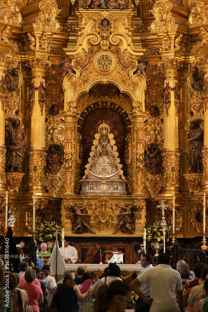 Faithful Catholics gathering around the altar of Ermita del Rocío church, a priest reading Sunday mass, the richly decorated sculpture of the Virgin with Child in the back, El Rocío, Spain