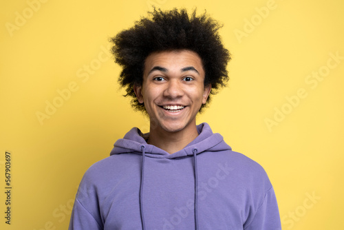 Attractive smiling African American guy wearing stylish purple hoodie isolated on yellow background, positive emotions. Portrait of young happy student looking at camera © Maria Vitkovska