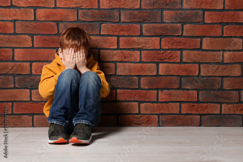 Upset boy sitting on floor near brick wall, space for text. Children's bullying photo