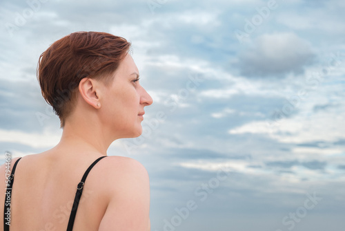 Portrait of a middle-aged woman in profile against a cloudy sky. A woman in a bathing suit is relaxing on the beach. A woman with short red hair looks away dreamily.
