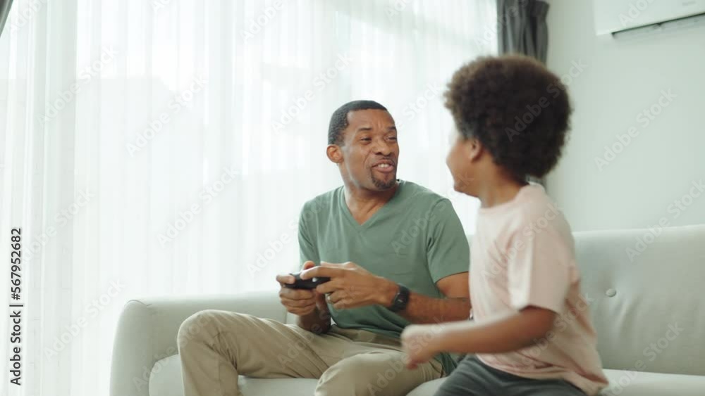Black African American Father and his little son sitting on the couch playing video games at home, Having fun.
Black African American Father and his little son sitting on the couch playing video games at home, Having fun.
