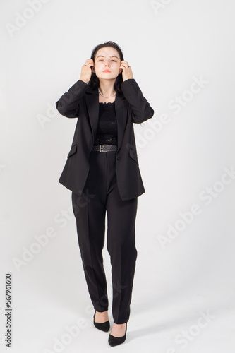 A young brunette woman poses standing in front of a white background / The model is dressed in a black jacket and trousers, there is a lot of free space around her for advertising