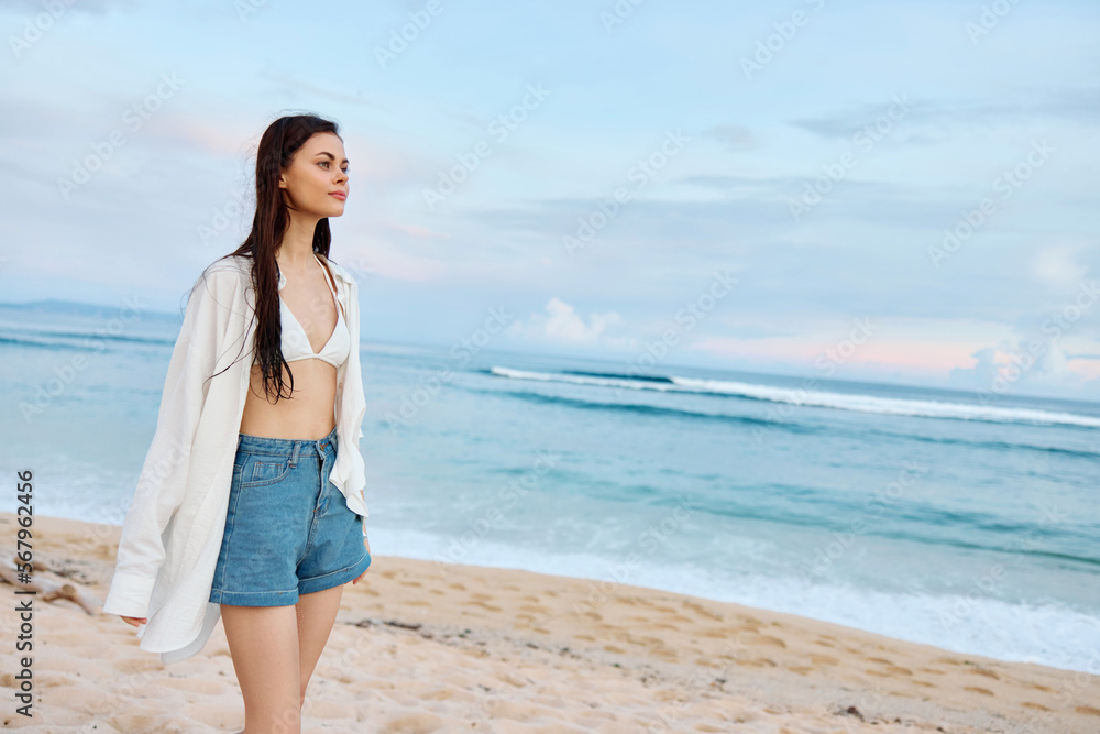 Happy tanned slim woman in white swimsuit shirt and denim shorts walks on the beach on the sand by the ocean with wet hair after swimming, sunset light in Bali Happy tanned slim woman in white swimsuit shirt and denim shorts walks on the beach on the sand by the ocean with wet hair after swimming, sunset light in Bali