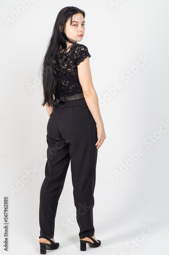 A young brunette poses from the back in half a turn on a white background / The model is dressed in a black jacket and trousers, there is a lot of free space around her for advertising