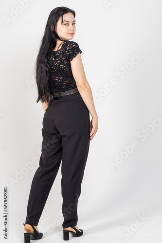 A young brunette poses from the back in half a turn on a white background / The model is dressed in a black jacket and trousers, there is a lot of free space around her for advertising