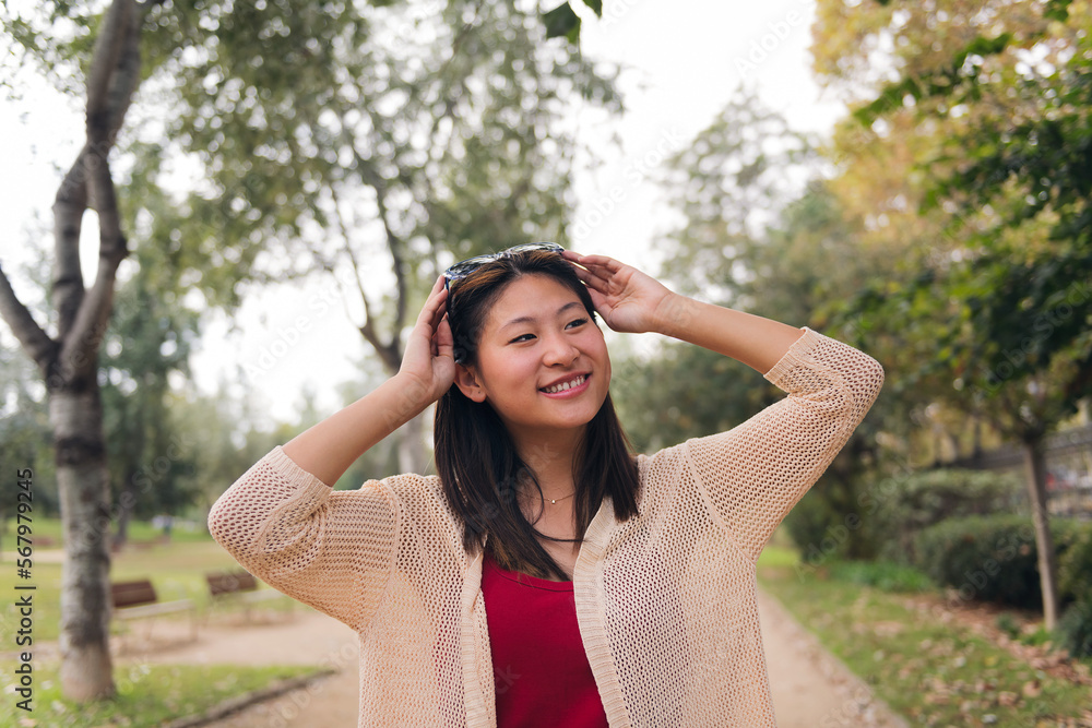 cheerful asian woman putting on her sunglasses on her head smiling in the park, concept of youth and beauty