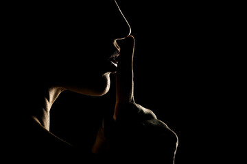 Silhouette of unknown woman with the face in the shadow holding finger on her lips on a black background