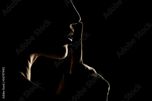 Silhouette of unknown woman with the face in the shadow holding finger on her lips on a black background