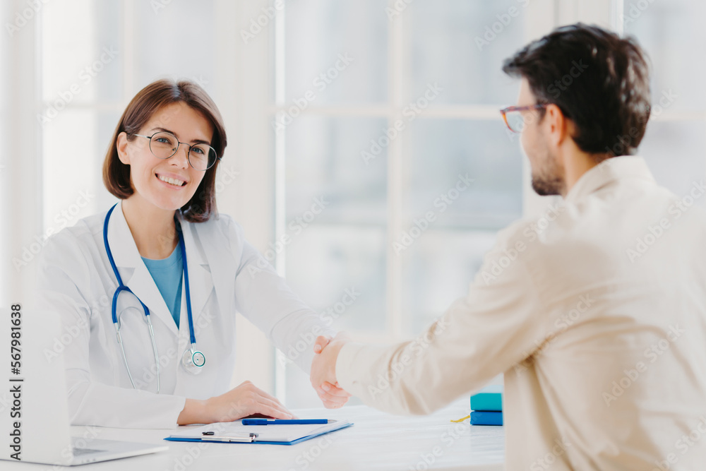 Partnership, assistance, trust and medicine concept. Female doctor shakes hands with thankful patient for good treatment and professionalism, pose in clinic, medical records near on white table Partnership, assistance, trust and medicine concept. Female doctor shakes hands with thankful patient for good treatment and professionalism, pose in clinic, medical records near on white table