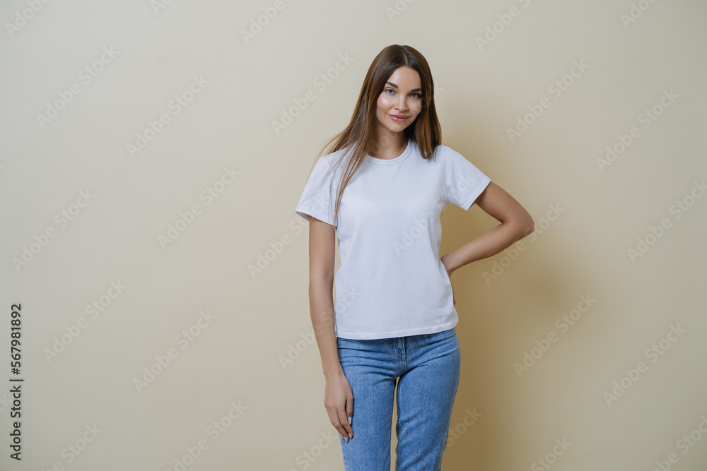 Attractive young female keeps hand on waist, wears casual clothes, ready for going out during weekend, enjoys nice weather, expresses joyful positive attitude to interlocutor, poses over beige wall Attractive young female keeps hand on waist, wears casual clothes, ready for going out during weekend, enjoys nice weather, expresses joyful positive attitude to interlocutor, poses over beige wall
