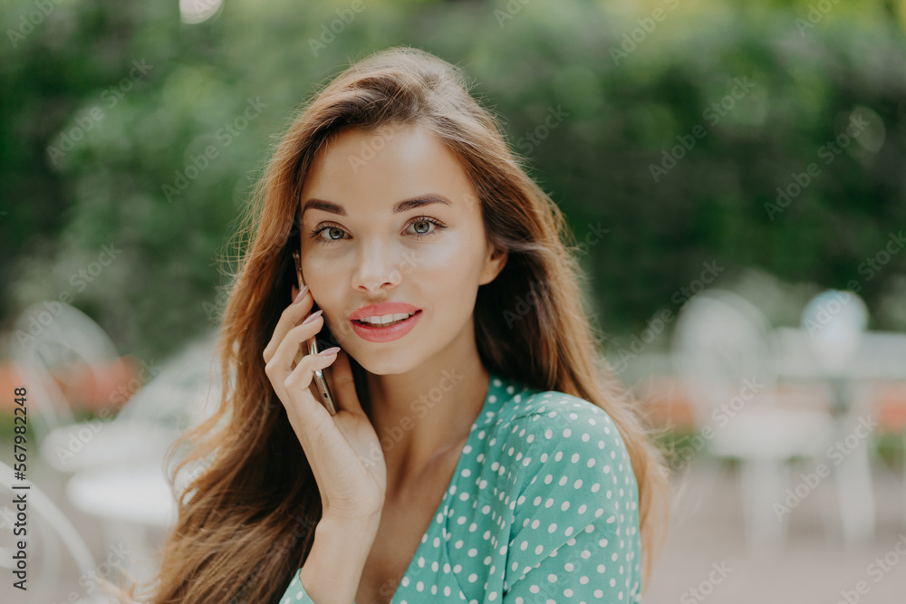 Horizontal shot of pretty young woman wears polkadot blouse, makes phone call, speaks with friend, poses outside against green blurred background, wears lipstick. People and communication concept Horizontal shot of pretty young woman wears polkadot blouse, makes phone call, speaks with friend, poses outside against green blurred background, wears lipstick. People and communication concept