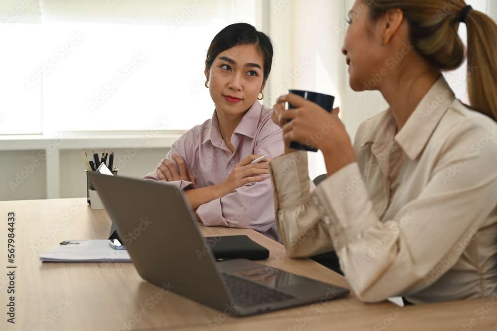 Two smiling women colleagues chatting, discussing team project during coffee break in modern office Two smiling women colleagues chatting, discussing team project during coffee break in modern office