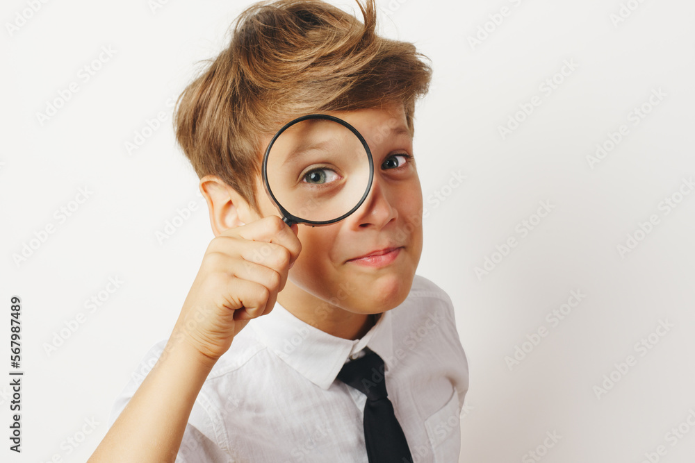 Funny schoolboy looks through a magnifying glass. Dressed in a white shirt and tie. Formal wear. White background, close-up studio portrait. Funny schoolboy looks through a magnifying glass. Dressed in a white shirt and tie. Formal wear. White background, close-up studio portrait.