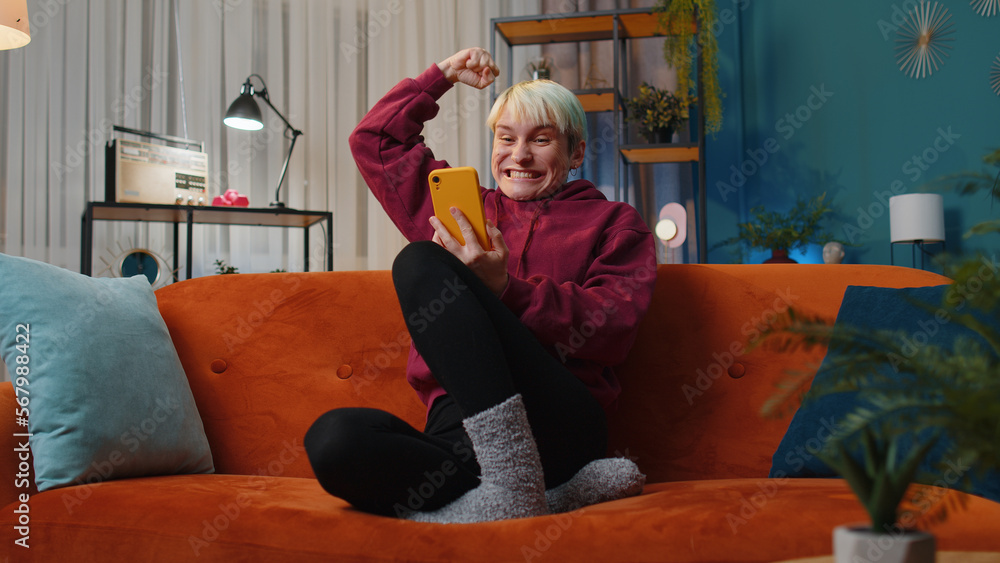 Obraz premium Happy woman with short hair use mobile smartphone typing browsing say Wow yes found out great big win good news celebrate lottery jackpot doing winner gesture. Girl at home in room sitting on sofa