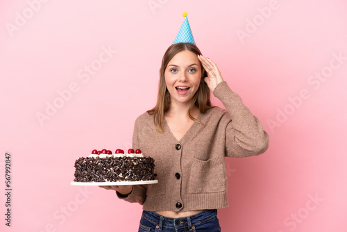 Young Lithuanian woman holding birthday cake isolated on pink background with surprise expression