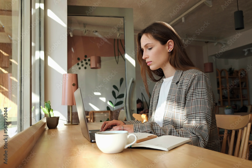 Beautiful young woman sitting in coffee shop with laptop and a planner. Female freelancer wearing smart casual outfit working in coffeehouse. Remote job concept. Close up, copy space, background. Beautiful young woman sitting in coffee shop with laptop and a planner. Female freelancer wearing smart casual outfit working in coffeehouse. Remote job concept. Close up, copy space, background.