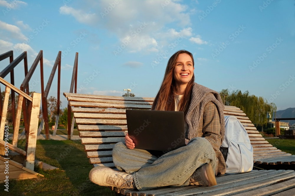 Young smiling woman sitting sitting with the laptop and working on the wooden deck chair in the park. Natural soft sunset light, copy space, background. Young smiling woman sitting sitting with the laptop and working on the wooden deck chair in the park. Natural soft sunset light, copy space, background.