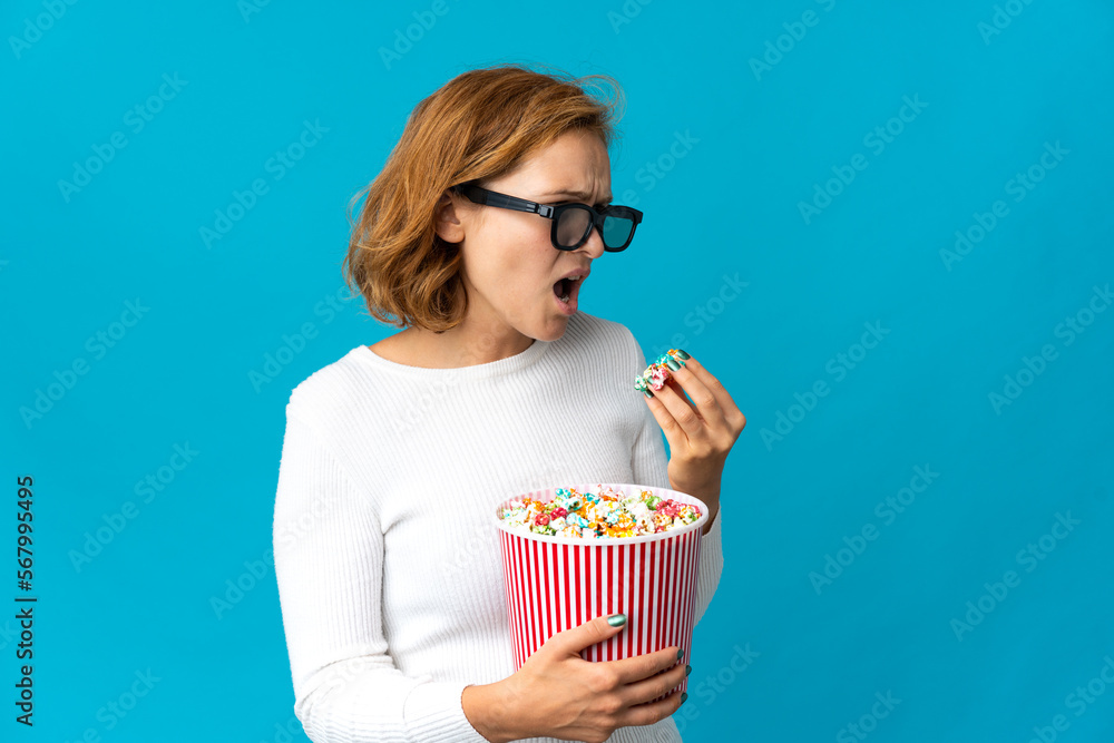 Young Georgian woman isolated on blue background with 3d glasses and holding a big bucket of popcorns while looking side