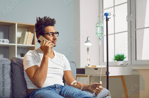 Young African American man talking with his doctor on his mobile phone while sitting on the sofa at home and receiving medication through an intravenous drip