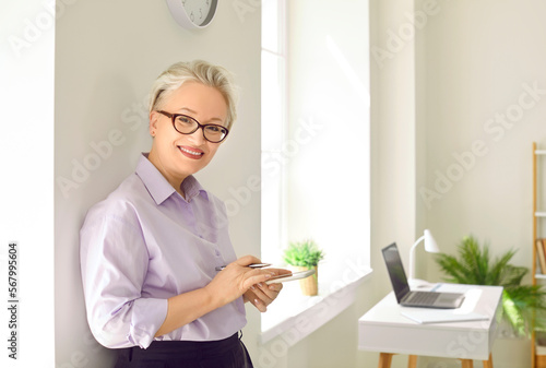 Successful elderly smiling woman senior business lady with mobile phone in hands looks at camera with confident look dressed in stylish formal clothes stand by window in own office