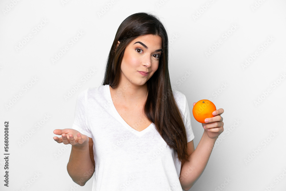 Young Brazilian woman holding an orange isolated on white background making doubts gesture while lifting the shoulders Young Brazilian woman holding an orange isolated on white background making doubts gesture while lifting the shoulders