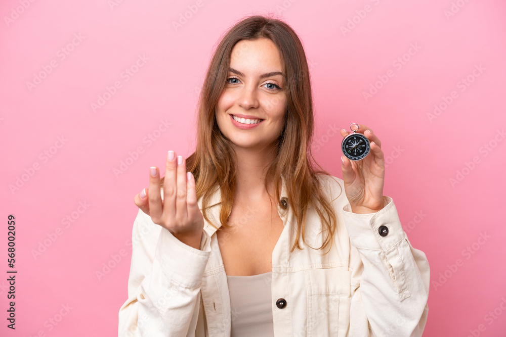 Young caucasian woman holding compass isolated on pink background inviting to come with hand. Happy that you came Young caucasian woman holding compass isolated on pink background inviting to come with hand. Happy that you came