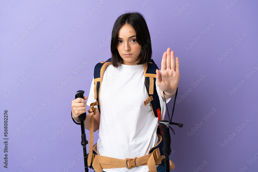 Young caucasian woman with backpack and trekking poles isolated on blue background making stop gesture