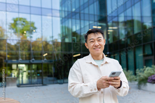 Portrait of successful Asian businessman in casual clothes, man with phone in hands smiling and looking at camera, business owner outside office building, satisfied freelancer startup entrepreneur. © Liubomir
