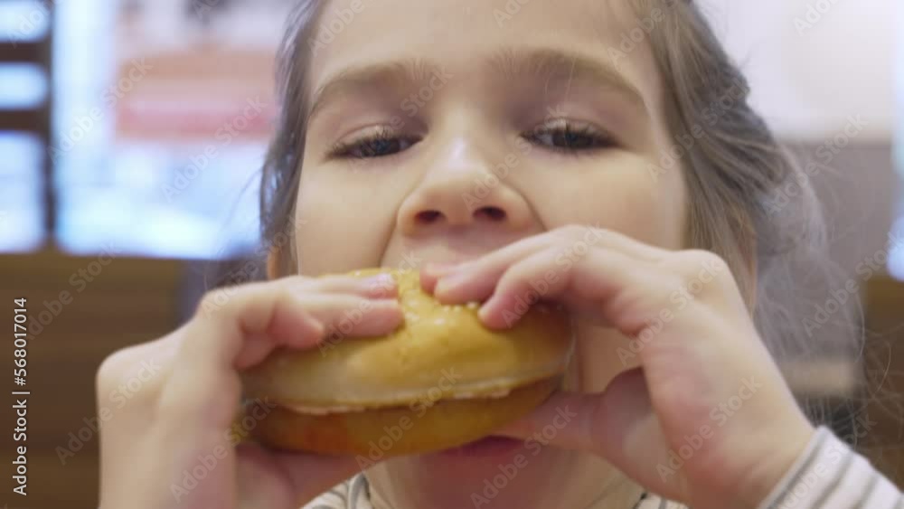 a cute little girl eats a burger in a cafe. the concept of fast food. harmful and tasty snack for children and adults. a cute little girl eats a burger in a cafe. the concept of fast food. harmful and tasty snack for children and adults.