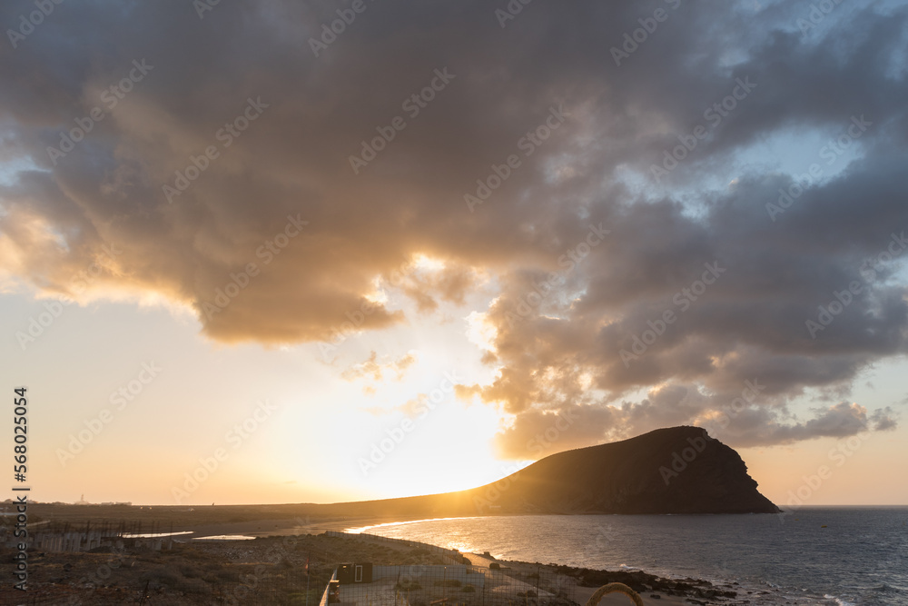 Sunset on the beach, golden sunbeams breaking through the gray storm clouds, with Red Mountain and ocen in the background. La Tejita, Tenerife, Canary Islands. Spain