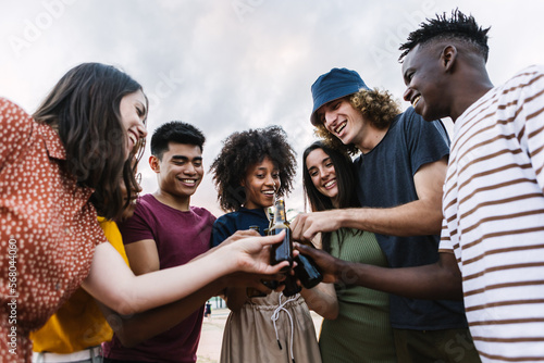 Young happy group of multi-ethnic friends celebrating together cheering with beer. Millennial diverse people enjoying summer party together on vacation. Friendship lifestyle concept