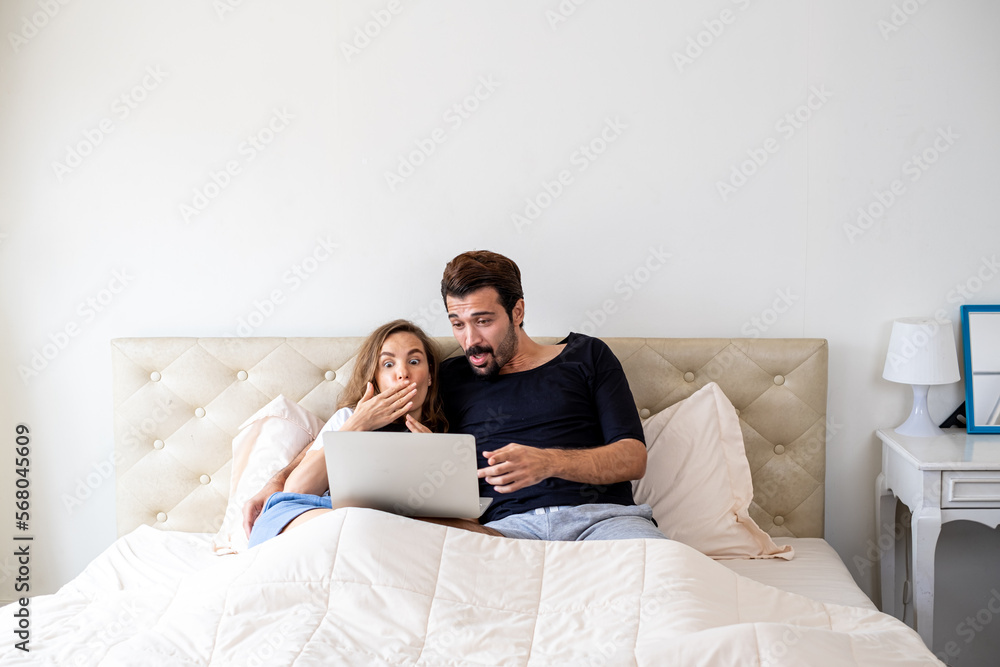 Fototapeta premium Happy Couple watching movie by laptop. Young couple watching a movie on bed at home in the afternoon. Man and woman watching movie from computer notebook on their bed.