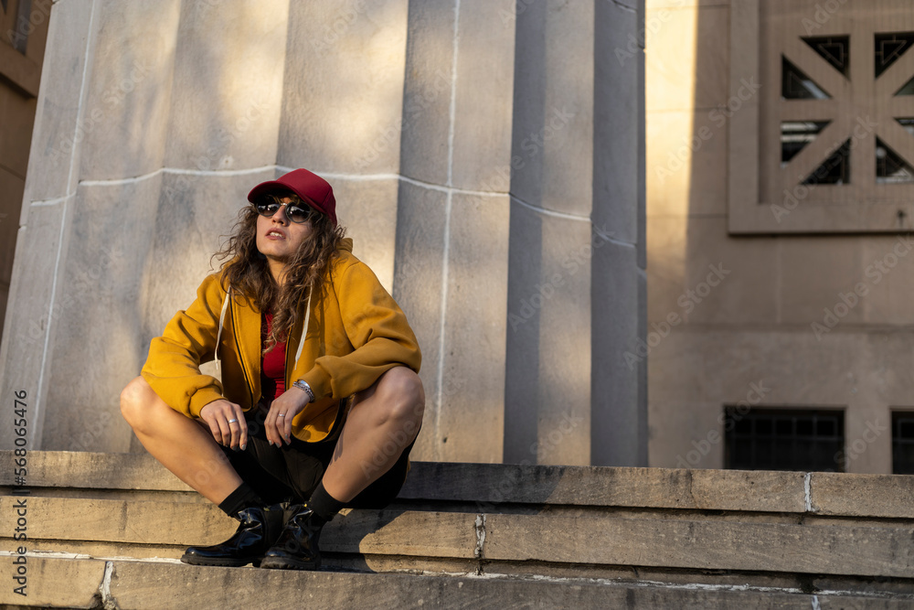 Latin American woman curly hair, red cap, sunglasses, sitting on stairs with relaxed attitude