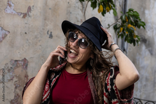Young Latin American woman with curly hair, hat, sunglasses, positive attitude talks on cell phone