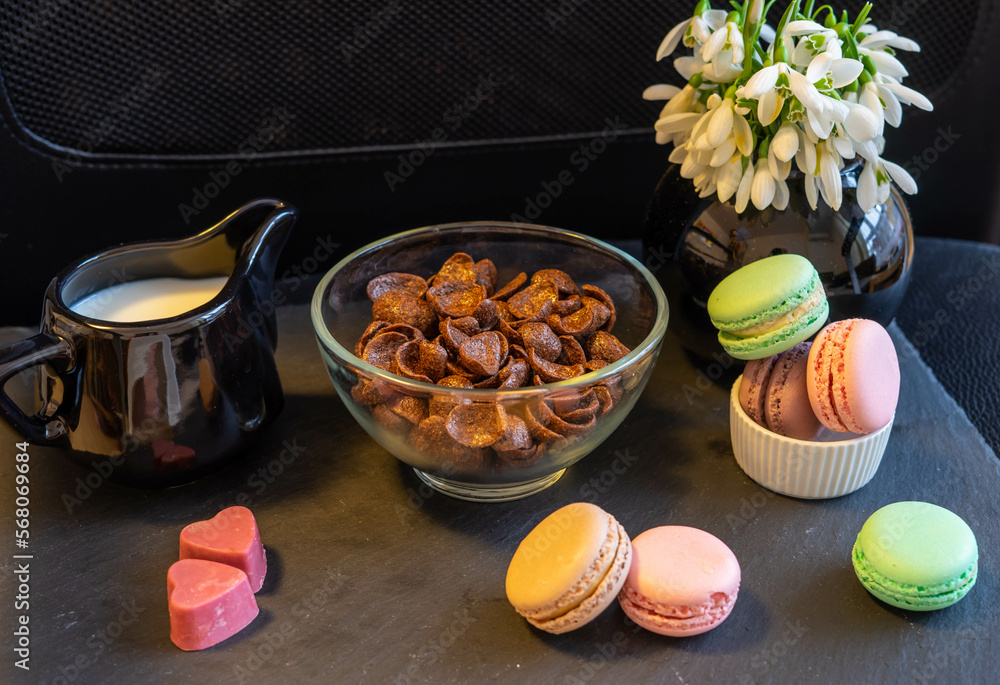 Breakfast with chocolate cereals, milk, macaroons, pink heart shaped chocolate pralines and snowdrops in a black vase, on a black background Breakfast with chocolate cereals, milk, macaroons, pink heart shaped chocolate pralines and snowdrops in a black vase, on a black background