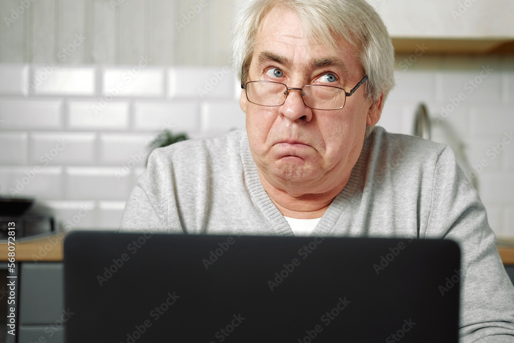 Senior grey haired man wearing glasses sitting at laptop and making fish face with lips. Crazy and comical gesture. Funny Old grandfather with playful silly facial expression grimacing fooling around Senior grey haired man wearing glasses sitting at laptop and making fish face with lips. Crazy and comical gesture. Funny Old grandfather with playful silly facial expression grimacing fooling around