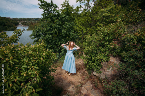A young girl with curly blond hair  in a blue dress  laughs and touches her hair with her hands  on a rock against the backdrop of trees and a picturesque river.