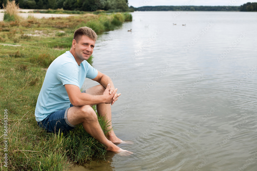 Fototapeta premium A smiling man with an athletic body, dressed in denim shorts and a blue t-shirt, sits on the banks of a picturesque river during the day.