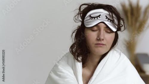 Sleepy young woman with a sleep mask on face, wrapped in blanket, falls on bed