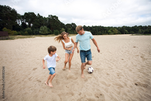 Family joint active leisure. Dad, mom and their little son play football on the sand, on the beach, by the river, against the backdrop of a picturesque landscape.