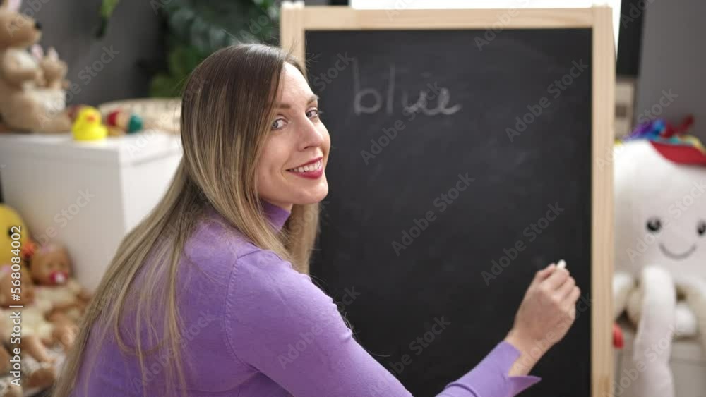 Young blonde woman preschool teacher writing blue word on blackboard at kindergarten Young blonde woman preschool teacher writing blue word on blackboard at kindergarten
