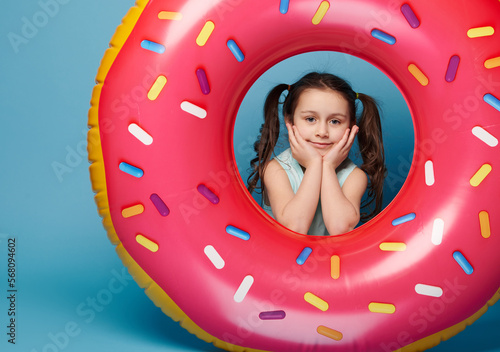 Beautiful little girl looking at camera through an inflatable colorful pink swim ring, isolated on blue color background