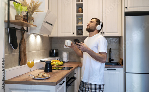 Man in headphones standing in domestic kitchen drinking coffee and listening music enjoying morning time.