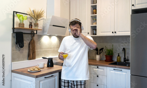 Sleepy young man in headphones standing in kitchen in morning with glass of orange juice and rubbing eyes with hands.