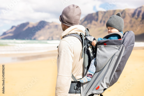 Young father carrying hisinf ant baby boy son in backpack on windy sandy Famara beach, Lanzarote island, Spain. Family travel and winter vacation concept © kasto