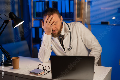 Male tired doctor with migraine overworked, overstressed working at night. sad unhappy health care professional with headache stressed sleepy photo